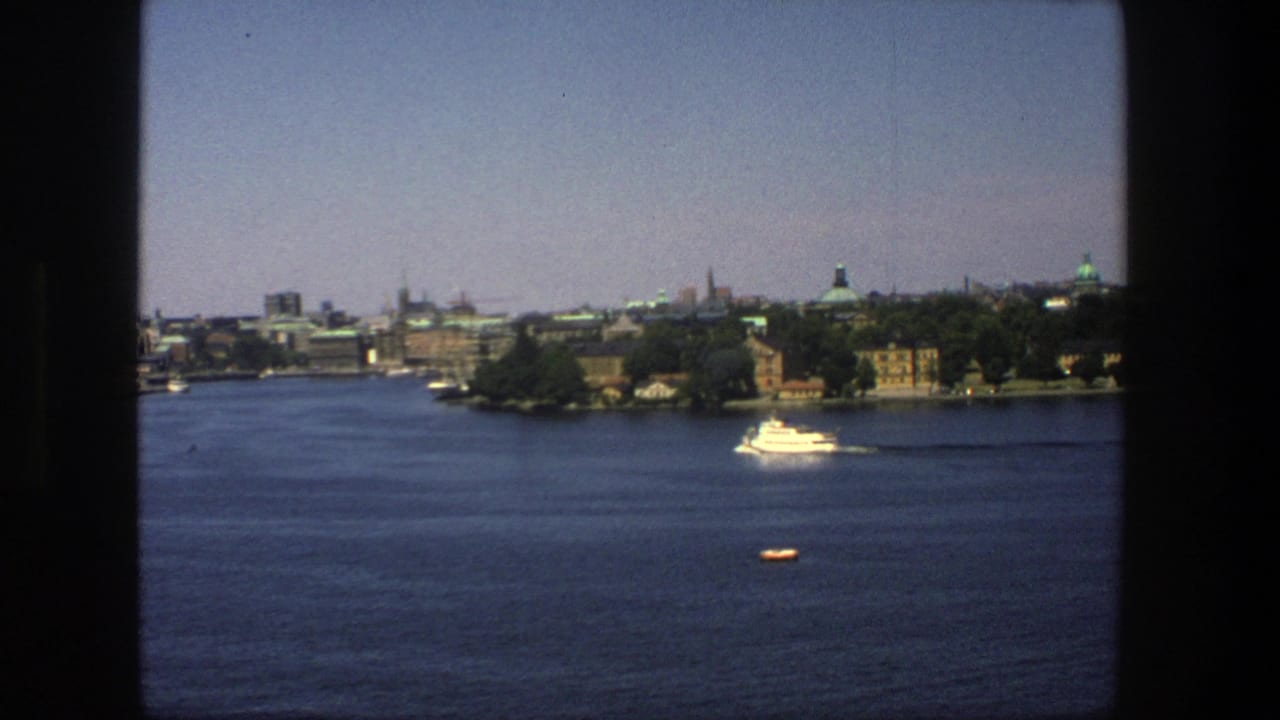 White passenger boat crossing a broad harbor with Stockholm buildings and trees in the distance.