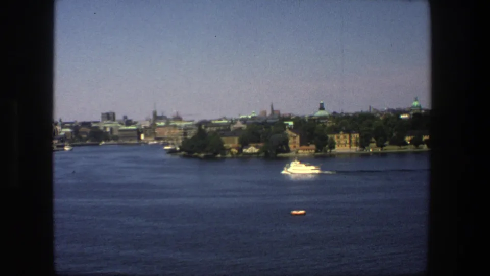 1975 Stockholm harbor view with a white passenger boat moving across blue water toward a distant tree-lined city shore.