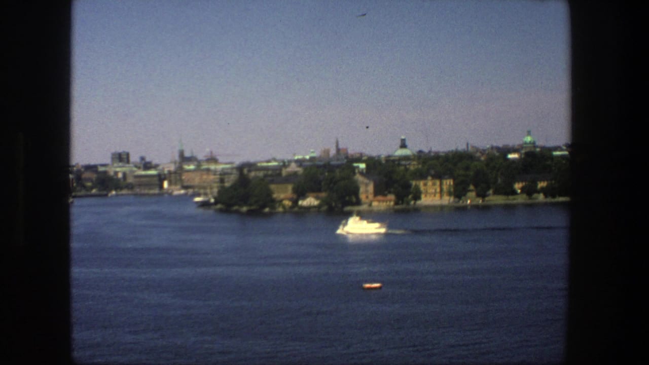 Stockholm waterfront seen from across the harbor, with a white passenger boat leaving a faint wake under a clear sky.