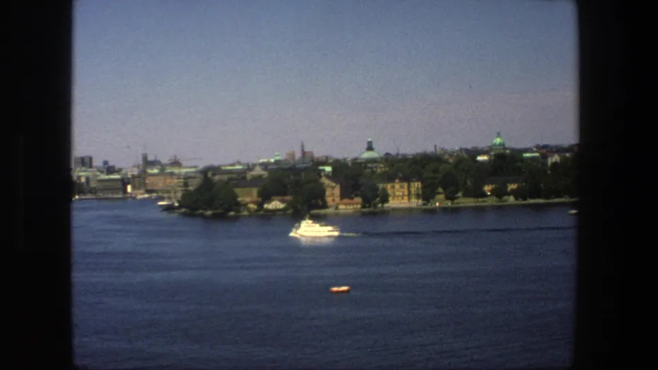 A visitor-shot 8mm frame shows a white boat crossing Stockholm harbor, with a small orange craft or buoy in the foreground water.