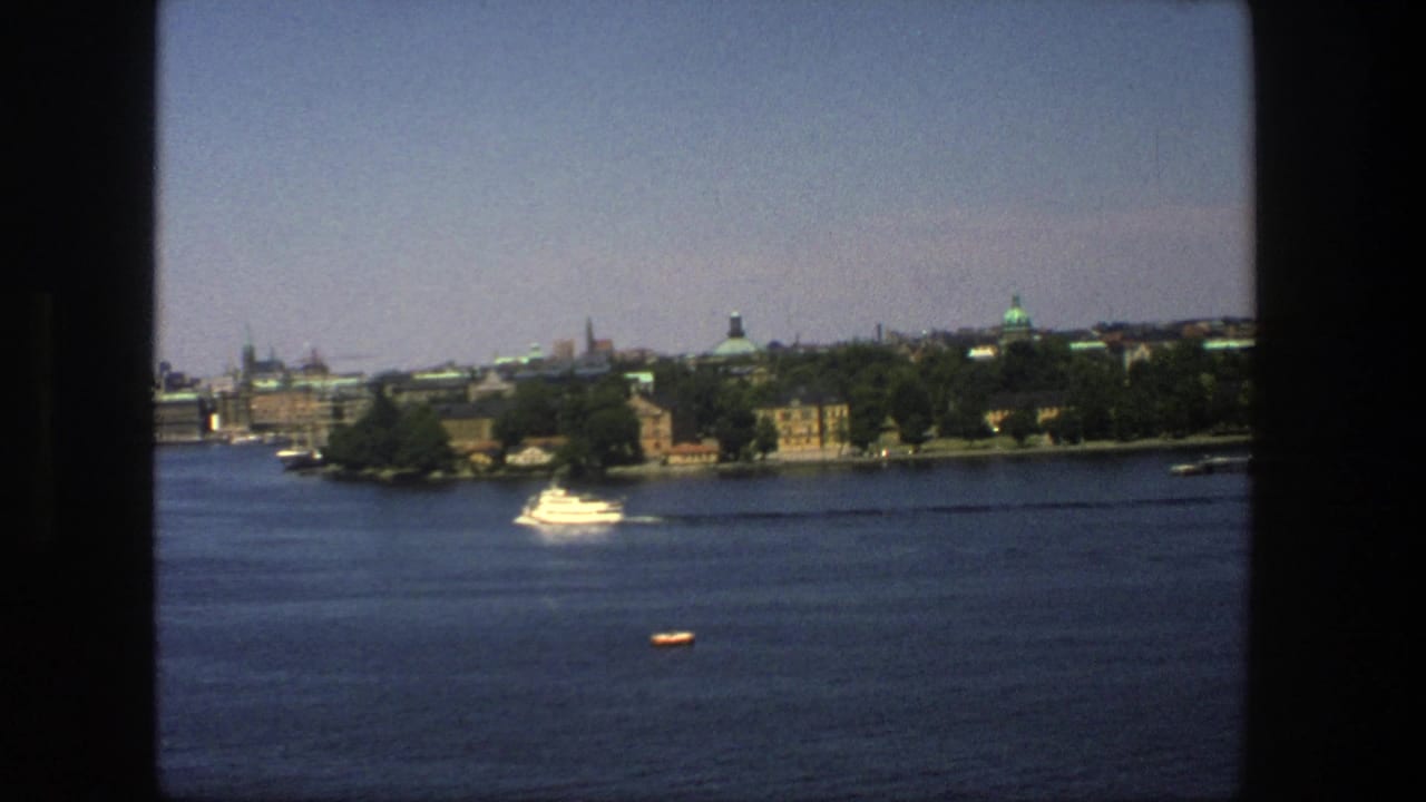 1975 8mm view of Stockholm from the water, with a white motorboat, tree-lined shore, and distant historic skyline.
