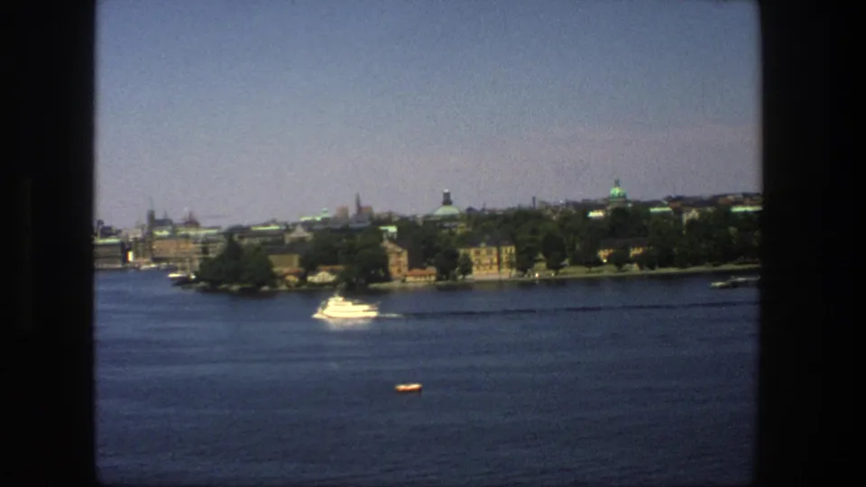 1975 8mm view of Stockholm from the water, with a white motorboat, tree-lined shore, and distant historic skyline.