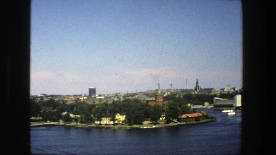 1975 8mm Stockholm view across the water toward a wooded shoreline, moored boats, modern blocks, and distant spires.