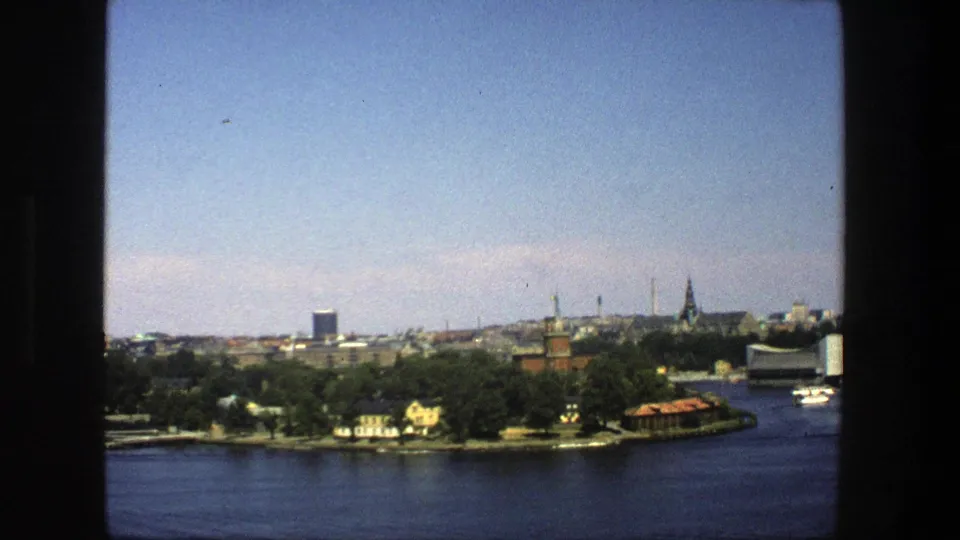 1975 Stockholm waterfront from the water, with a tree-lined shore, small boats, a red-roofed building, and distant city towers.