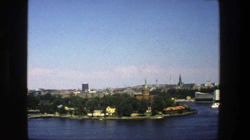 Vintage 8mm Stockholm panorama showing water in the foreground, trees along the shore, boats, towers, and historic rooflines.