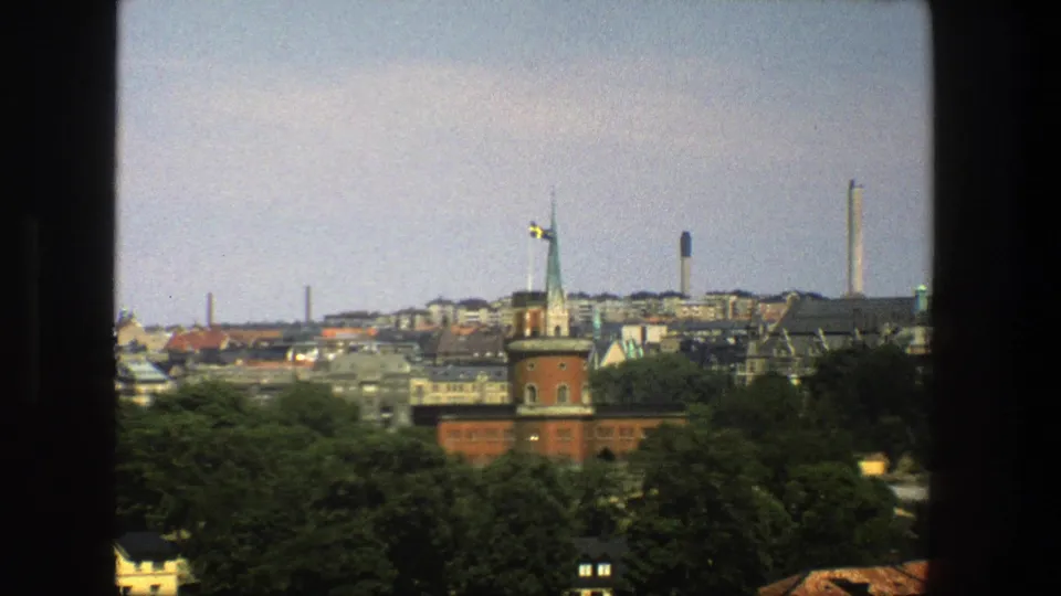 1975 8mm close view of Stockholm architecture with a red-brick spire, Swedish flag, treetops, rooftops, and modern towers.