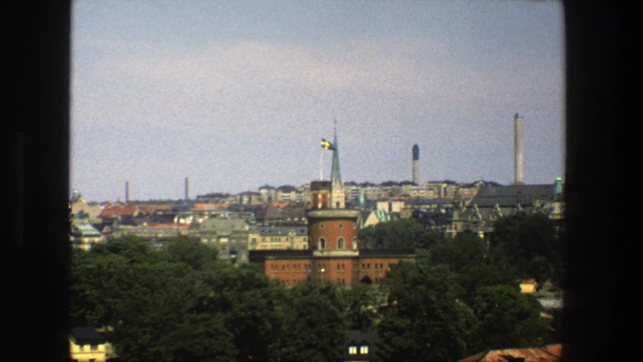 1975 8mm view of Stockholm through a window, showing a red-brick tower, Swedish flag, treetops, rooftops, and distant smokestacks.