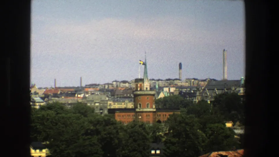 1975 8mm view of Stockholm through a window, showing a red-brick tower, Swedish flag, treetops, rooftops, and distant smokestacks.