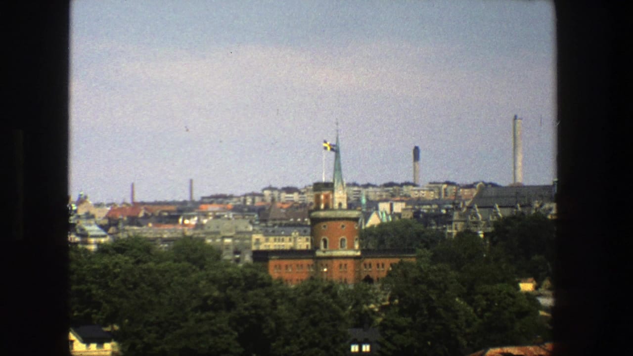 1975 Stockholm city view from behind a window frame, with a red-brick tower, Swedish flag, dense trees, rooftops, and industrial chimneys.