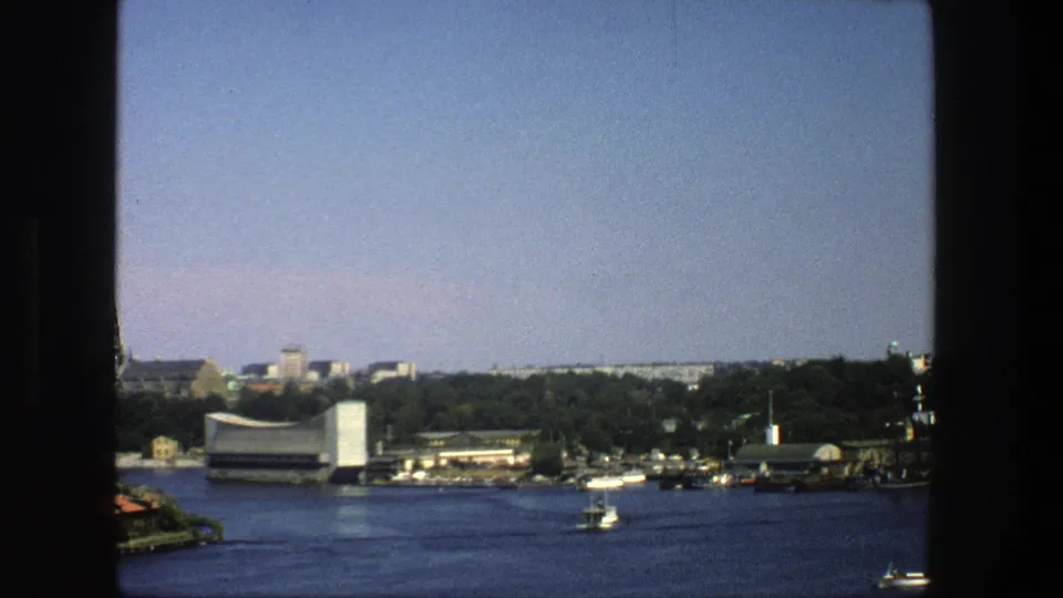 1975 Stockholm waterfront view showing open blue water, small boats, tree-lined shore, low buildings, and a modern white waterfront structure.