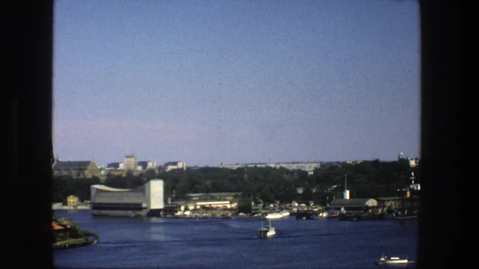 1975 8mm Stockholm harbor view with blue water, small boats, a wooded shoreline, low buildings, and a pale sky during the pan.