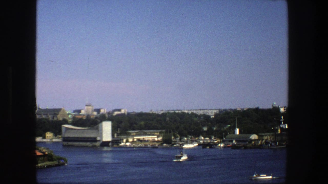 1975 8mm view across Stockholm water toward shoreline buildings, trees, and small boats under a clear sky.