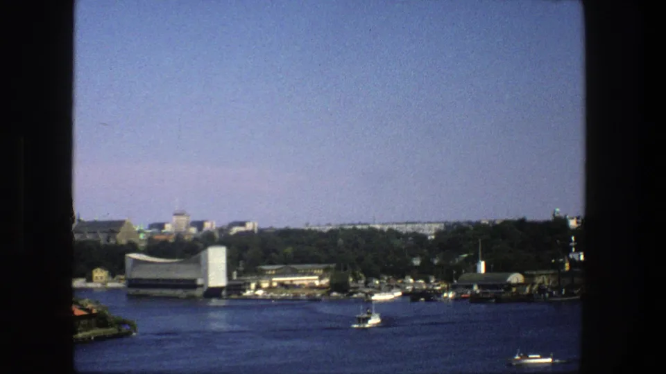 1975 8mm view across Stockholm water toward shoreline buildings, trees, and small boats under a clear sky.
