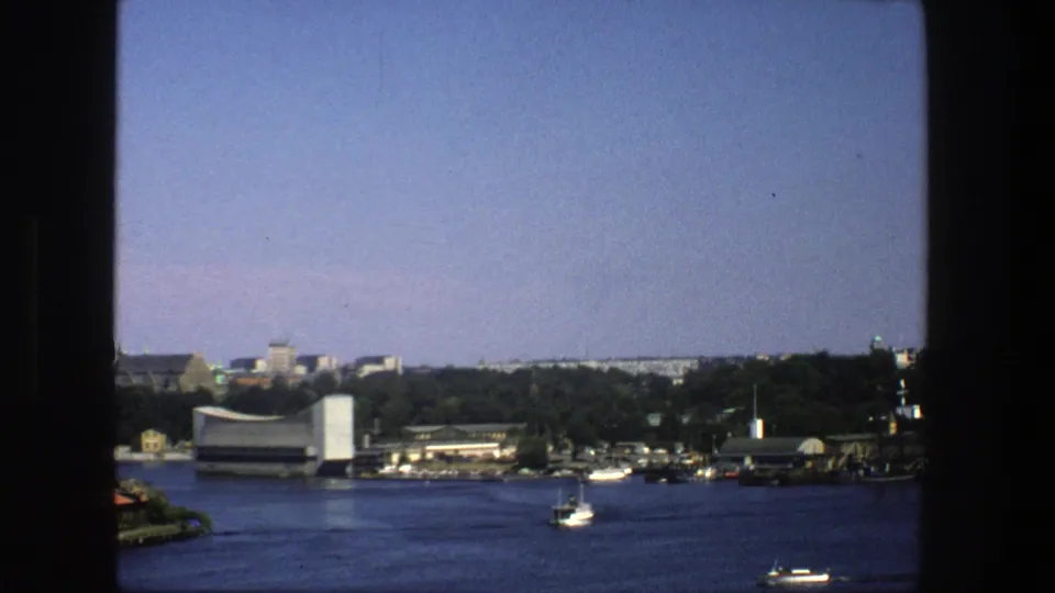 Stockholm waterfront seen from across the water in 1975, with small boats, trees, and low urban buildings visible.