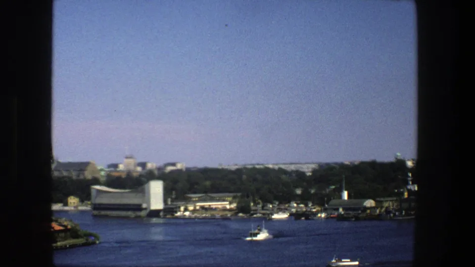 Soft 8mm frame of Stockholm water and shoreline, with a small boat passing below low buildings and trees.