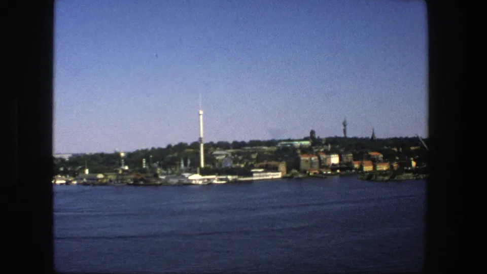 Later 1975 Stockholm waterfront view showing a tall tower, moored or moving boats, and a darker tree-lined shore.