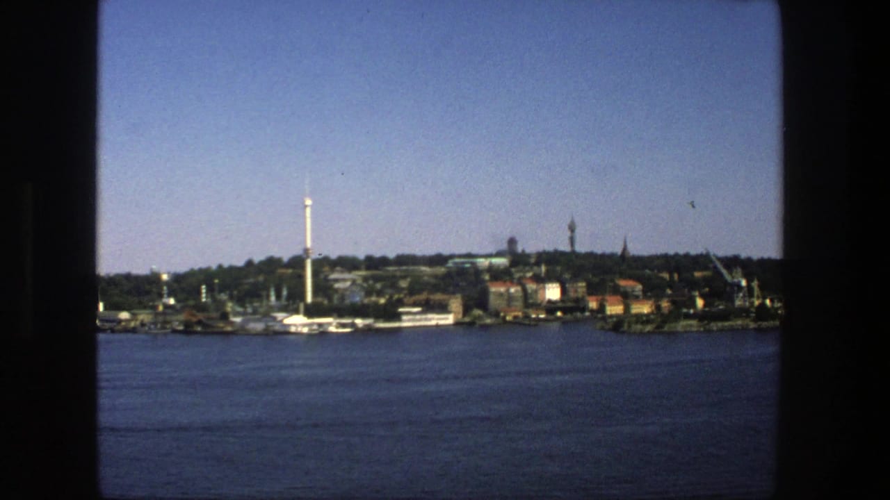 Stockholm, Sweden in 1975, viewed from across the water with low waterfront structures, trees, cranes, and a prominent tower.