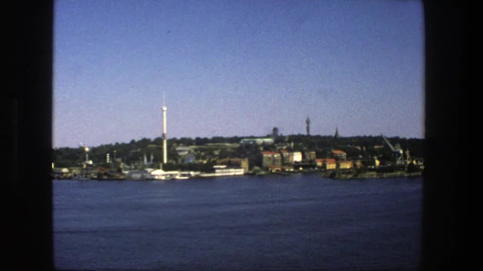 1975 home-movie frame of Stockholm across a body of water, showing docks, waterfront buildings, trees, and a tall tower under clear sky.