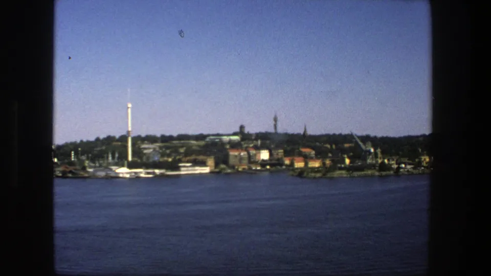Stockholm waterfront in 1975, with calm water in the foreground and an industrial shoreline marked by cranes, buildings, trees, and a tower.