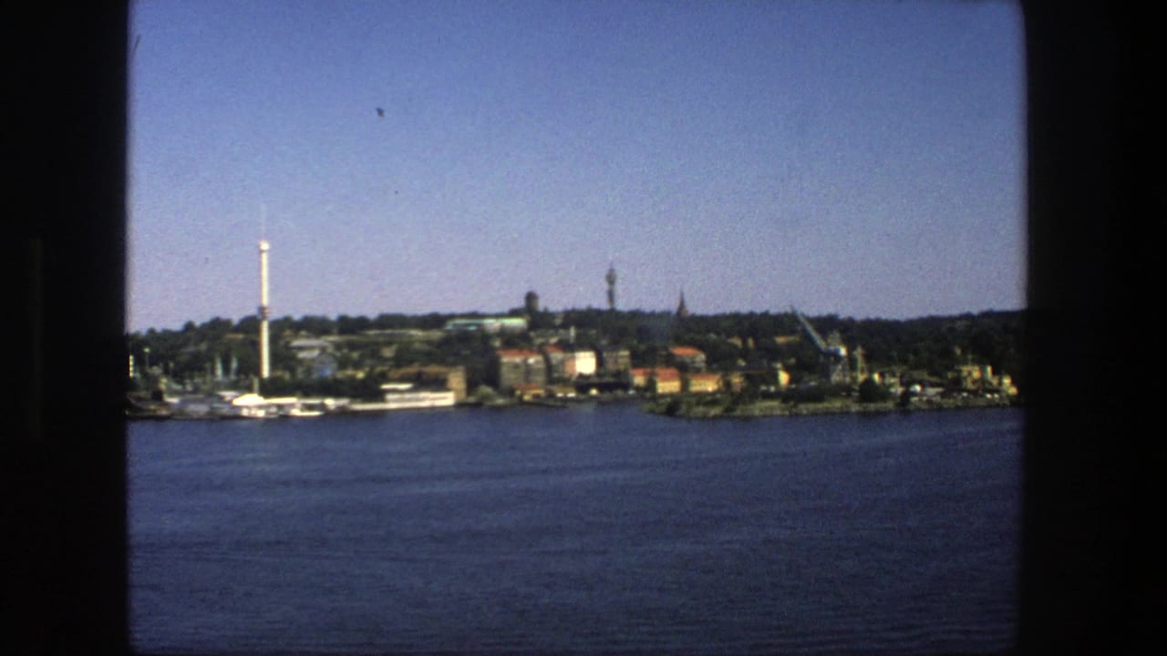 Distant Stockholm shoreline across blue water with buildings, trees, cranes, and a tall white tower.