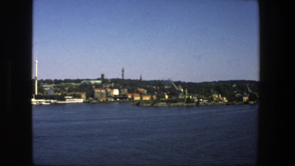 1975 8mm Stockholm shoreline scene with blue water in the foreground and distant buildings, cranes, trees, and towers beyond.
