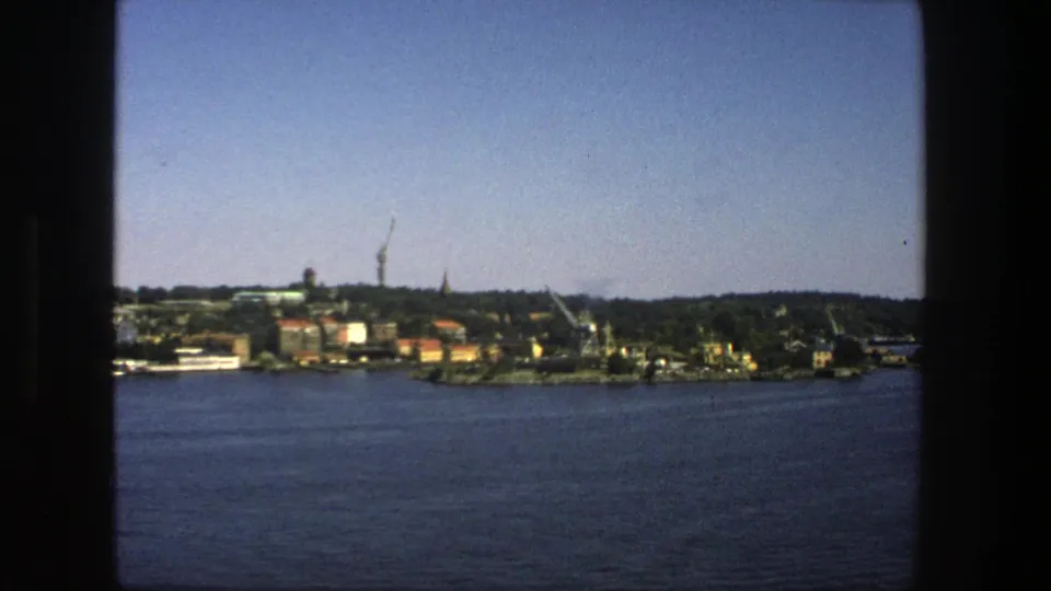 1975 Stockholm home-movie frame showing a distant urban shoreline across water, with trees, buildings, cranes, and a spire under open sky.