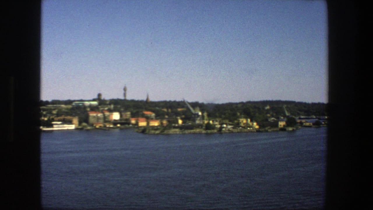 1975 8mm view from a boat toward Stockholm waterfront buildings, trees, cranes, and a tower across the water.