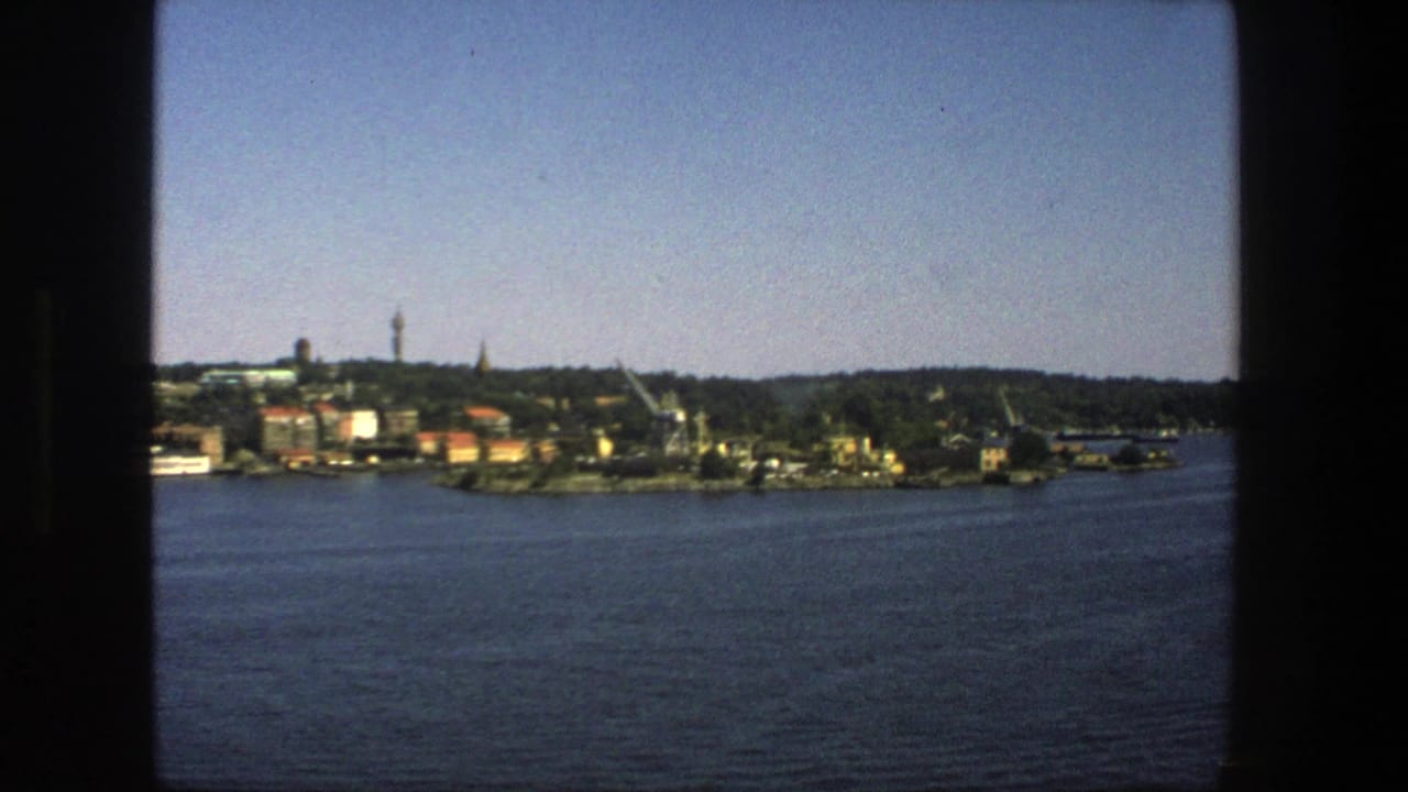 Stockholm waterfront appears across a broad stretch of water, with trees, buildings, cranes, and a tower visible in soft 8mm color.