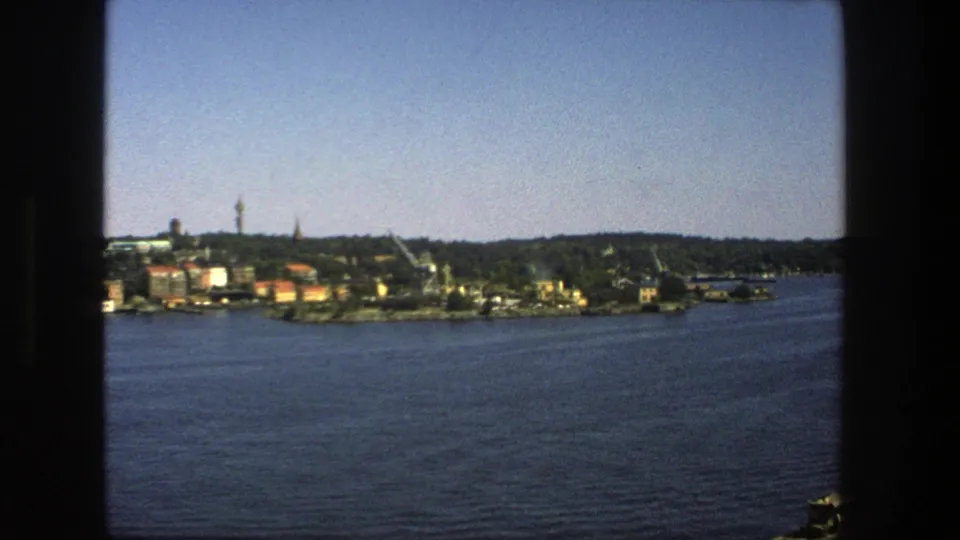 A 1975 home-movie frame shows Stockholm across the water, with shoreline structures, trees, cranes, and a prominent tower under clear sky.