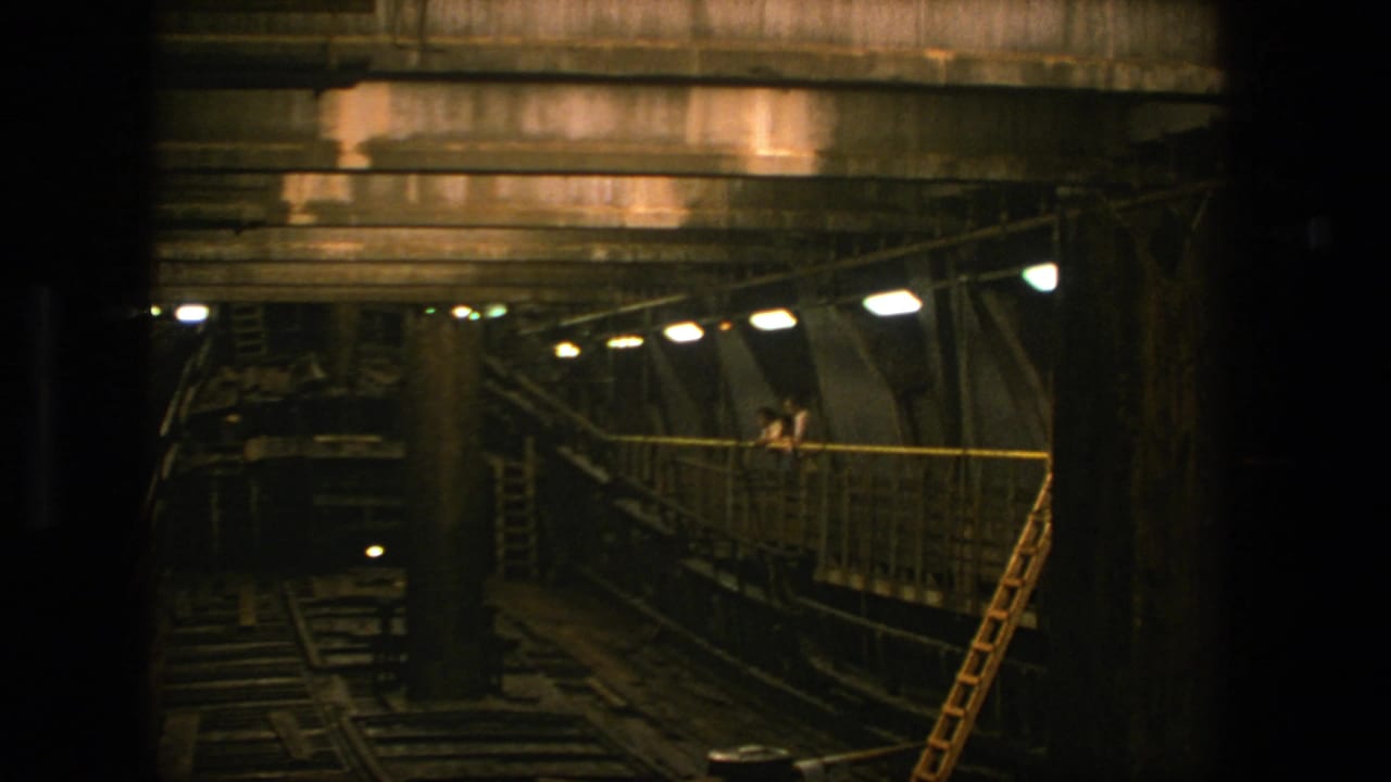 1975 Stockholm 8mm frame showing a dim industrial space with rails, overhead concrete rafters, a lit walkway, and a yellow ladder.