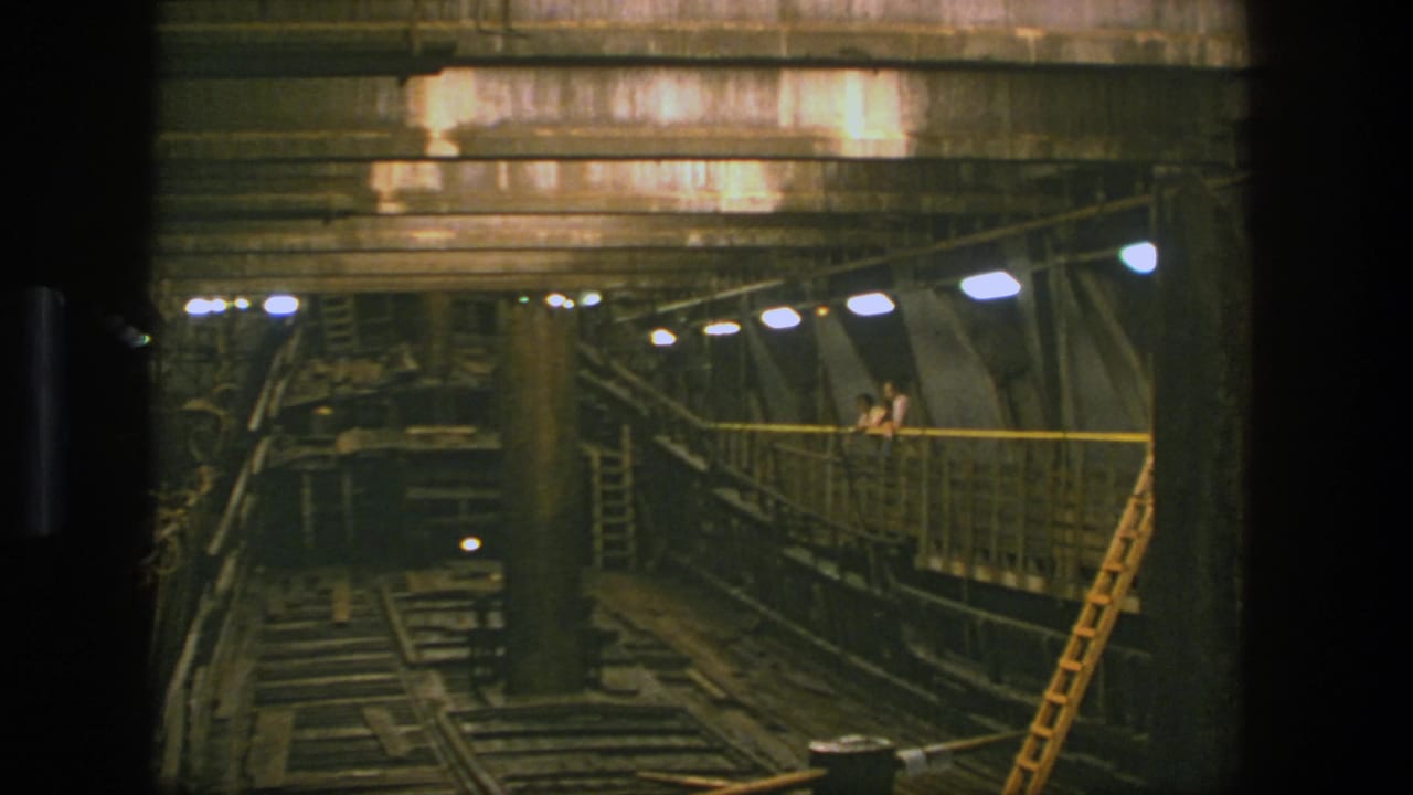1975 Stockholm frame of a low-lit industrial interior with rail tracks below and small people standing on a raised walkway.