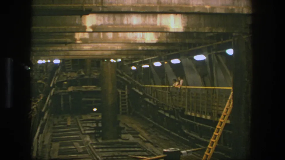 1975 Stockholm frame of a low-lit industrial interior with rail tracks below and small people standing on a raised walkway.