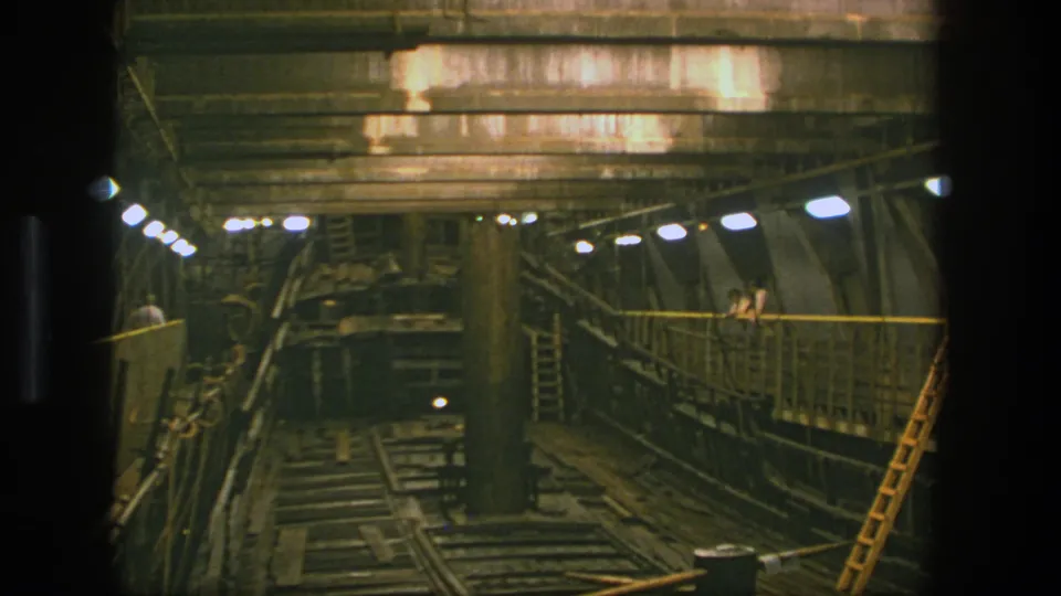 Wide 1975 Stockholm 8mm view of an industrial interior with tracks, lit walkway, concrete beams, and maintenance ladder.