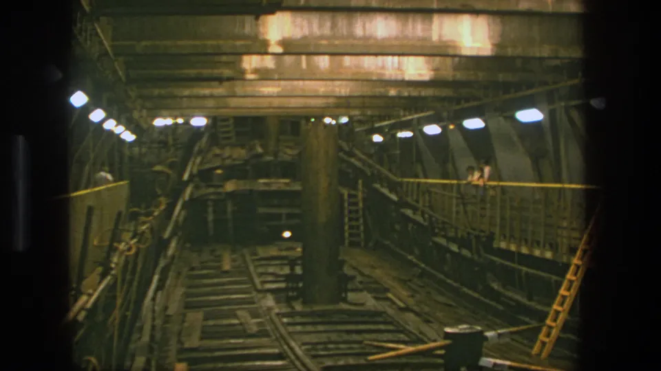 1975 Stockholm frame showing a dim rail-lined industrial interior, overhead concrete structure, walkway lights, and a yellow ladder.