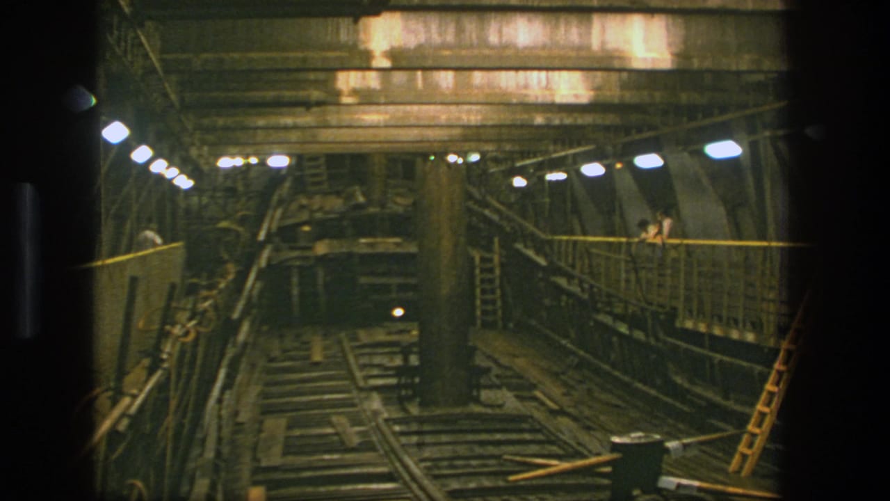 Dim shipyard interior with rails, timber supports, ladders, overhead lamps, and distant workers.