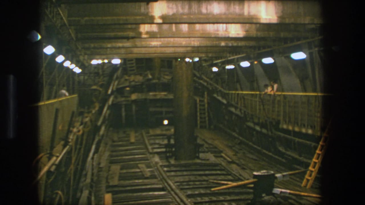 Industrial interior with overhead lamps, rails, ladders, walkways, and workers near a large metal structure.