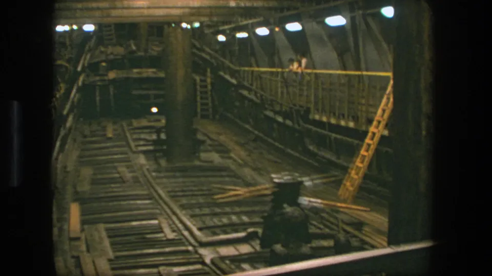 1975 Stockholm shipyard interior viewed from above, with rails, timber planks, metal walls, a leaning ladder, lamps, and workers in the background.