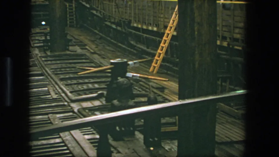 1975 Stockholm vessel construction interior with foreground railing, rail tracks, timber supports, a ladder, overhead lamps, and metal framing.