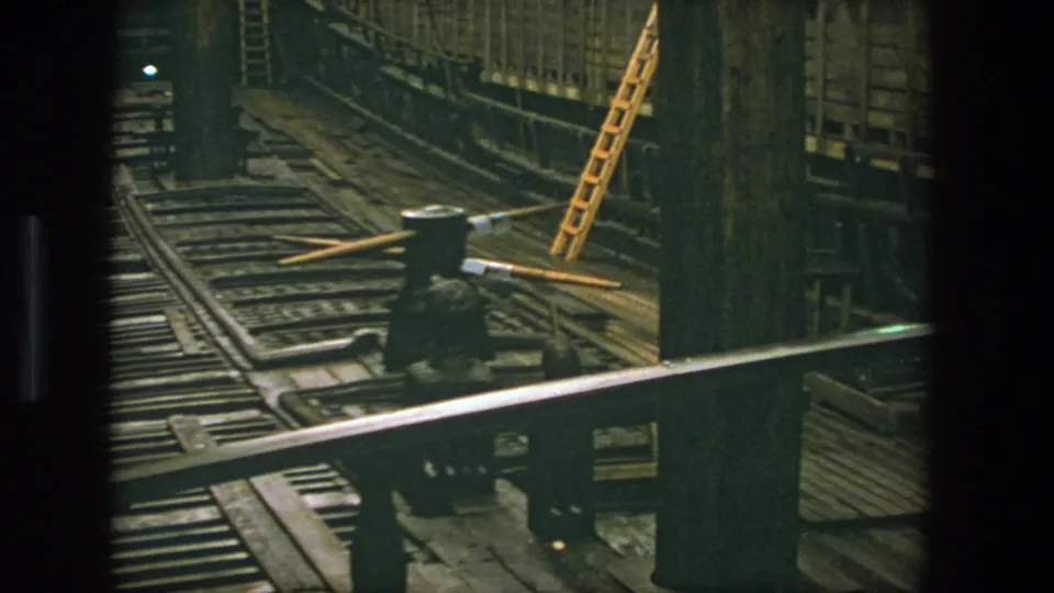 1975 Stockholm 8mm frame showing a dim industrial tunnel with rail tracks, heavy wood structure, a metal railing, and a yellow ladder.