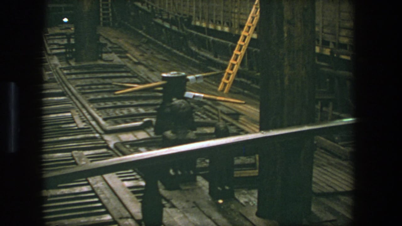 1975 Stockholm 8mm frame looking across rail tracks, timber framing, a metal railing, and a yellow ladder inside a dark industrial tunnel.