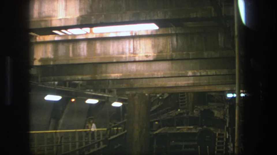 1975 Stockholm 8mm frame with a distant man walking on a raised metal-railed walkway in a shadowed industrial tunnel.