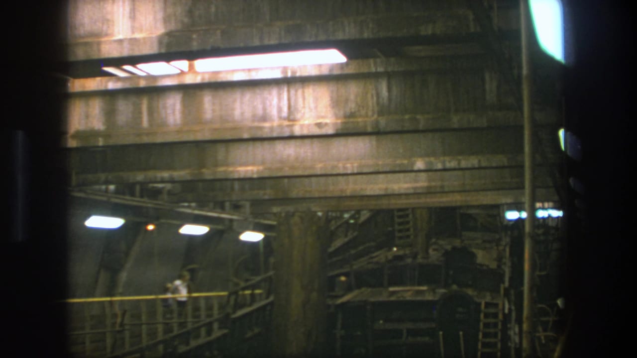 1975 Stockholm 8mm frame with a worker near railings inside a concrete worksite interior under fluorescent lighting.
