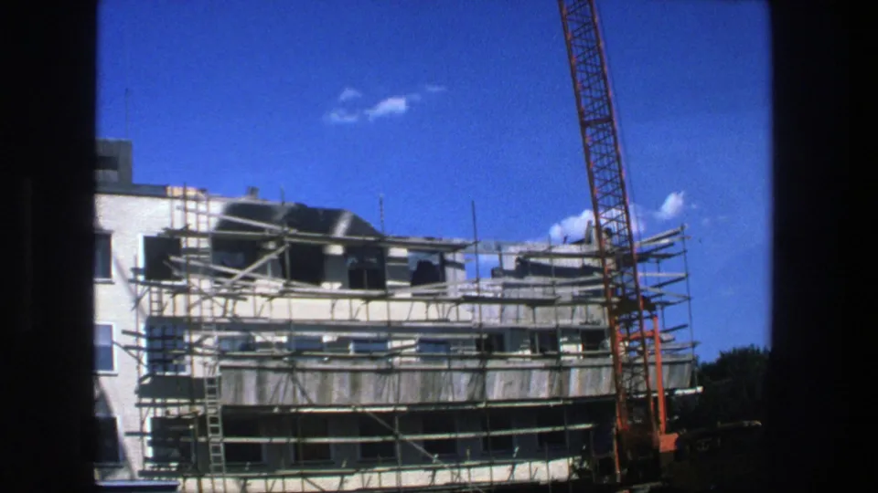 1975 Stockholm 8mm frame showing a scaffolded multi-story building and orange crane seen through dark window edges.