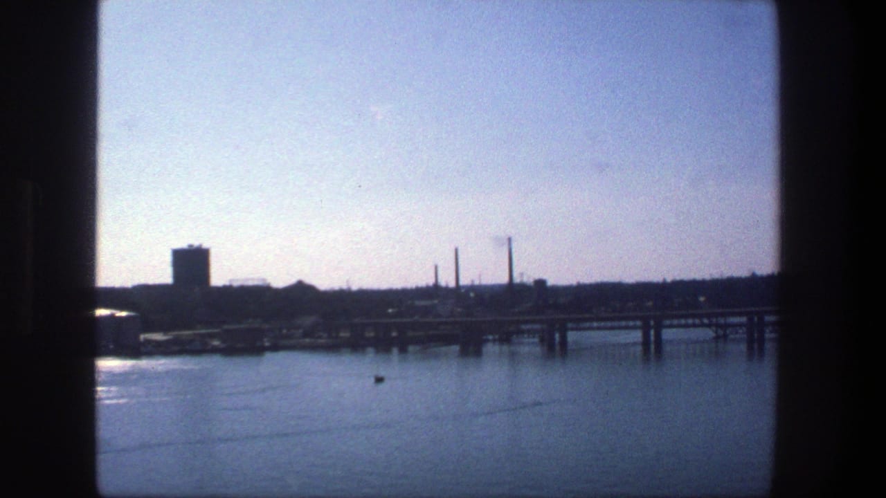 1975 Stockholm waterfront view with calm water, a docked cargo ship, bridge span, industrial buildings, and smokestacks in the distance.