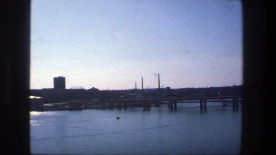 1975 Stockholm waterfront view with calm water, a docked cargo ship, bridge span, industrial buildings, and smokestacks in the distance.