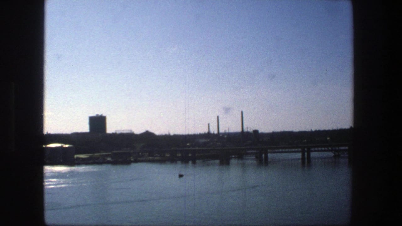 Stockholm harbor in 1975, seen across calm water toward a docked cargo vessel, bridge structure, industrial buildings, and smokestacks.