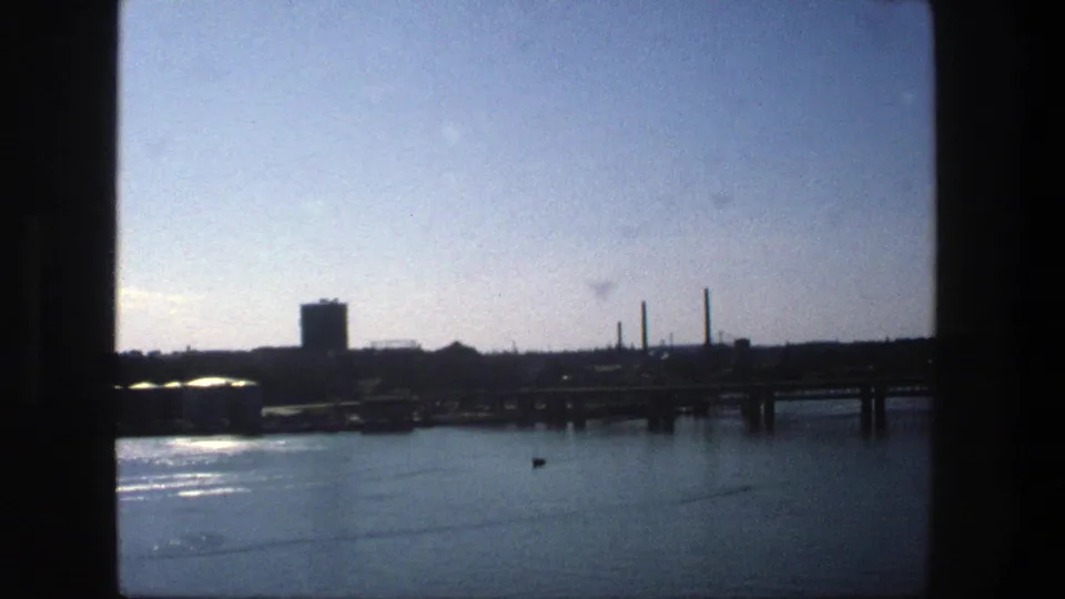 A 1975 Stockholm waterfront frame shows still harbor water, a distant docked ship, bridge or pier structure, and industrial shoreline.