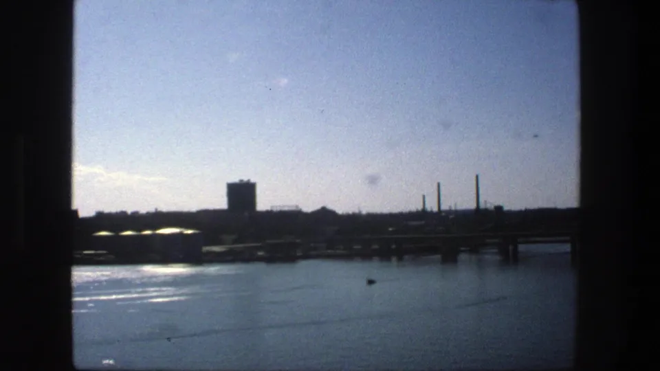 Final 1975 Stockholm harbor frame with reflected light on calm water, a docked cargo ship, bridge structure, and distant smokestacks.