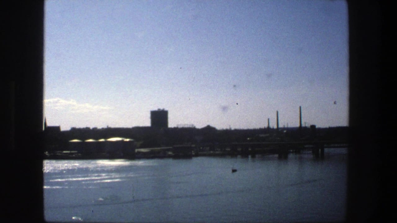 Waterfront view with distant industrial skyline, tower, smokestacks, and dark window edges.