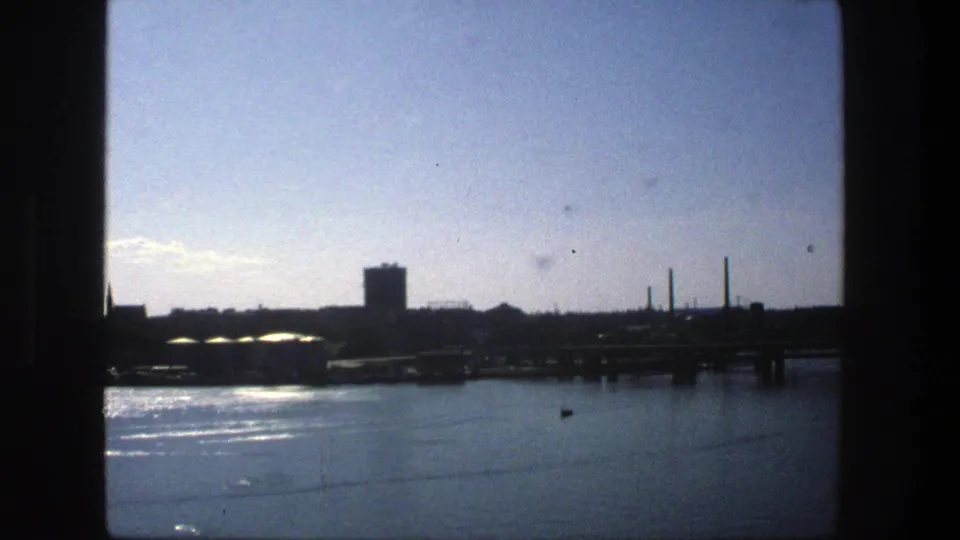 1975 Stockholm waterfront seen across calm water, with silhouetted industrial buildings, chimneys, a tower, and bright sky.